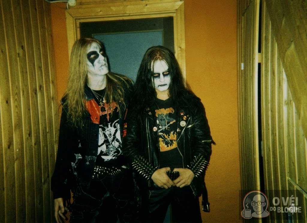 Fotografia de Dead e Euronymous da banda de Black Metal norueguesa Mayhem em um corredor de madeira. Ambos usam corpse paint, jaquetas de couro e camisetas de bandas de metal extremo.