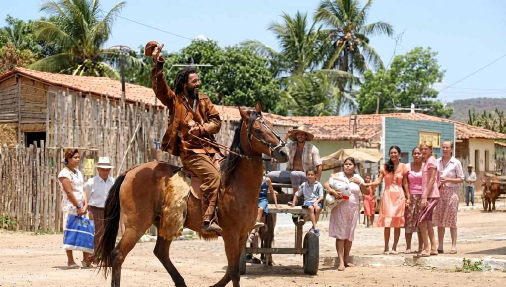 Close do ator Marcos Palmeira como Ojuara, sorrindo e usando chapéu de couro sertanejo, em cena do filme O Homem que Desafiou o Diabo.